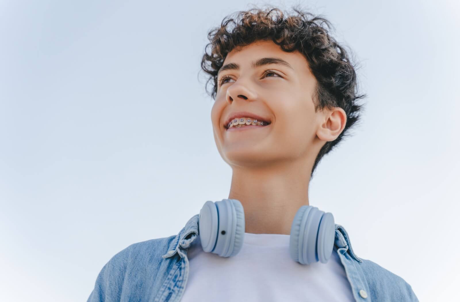 Smiling teenager with braces wearing a denim jacket and headphones around their neck, looking off into the distance against a clear blue sky.